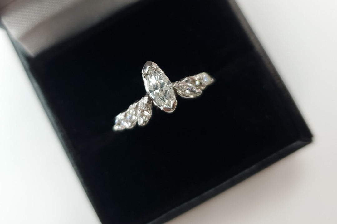 A white gold ring with a marquise-cut clear center diamond and smaller clear stones along the band, displayed on a black ring box against a white background.